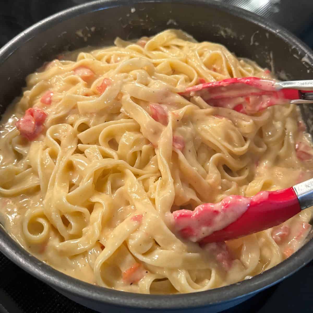 skillet with tongs with he making creamy fettuccine noodles